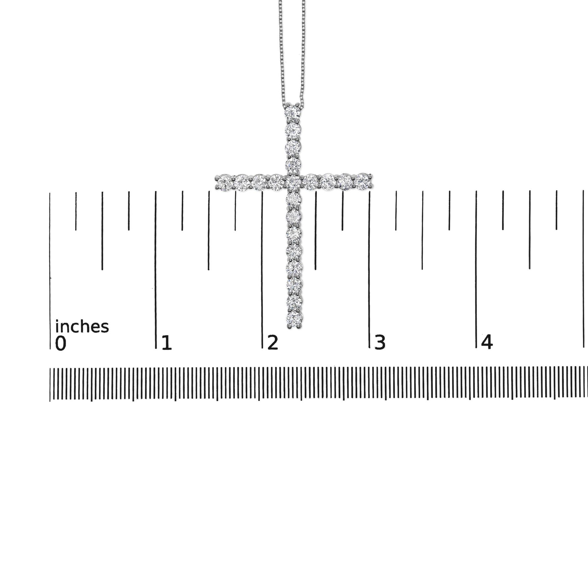 Close-up front view of a white metal cross necklace pendant with round white gemstones in prong settings, shown on a ruler for size reference.