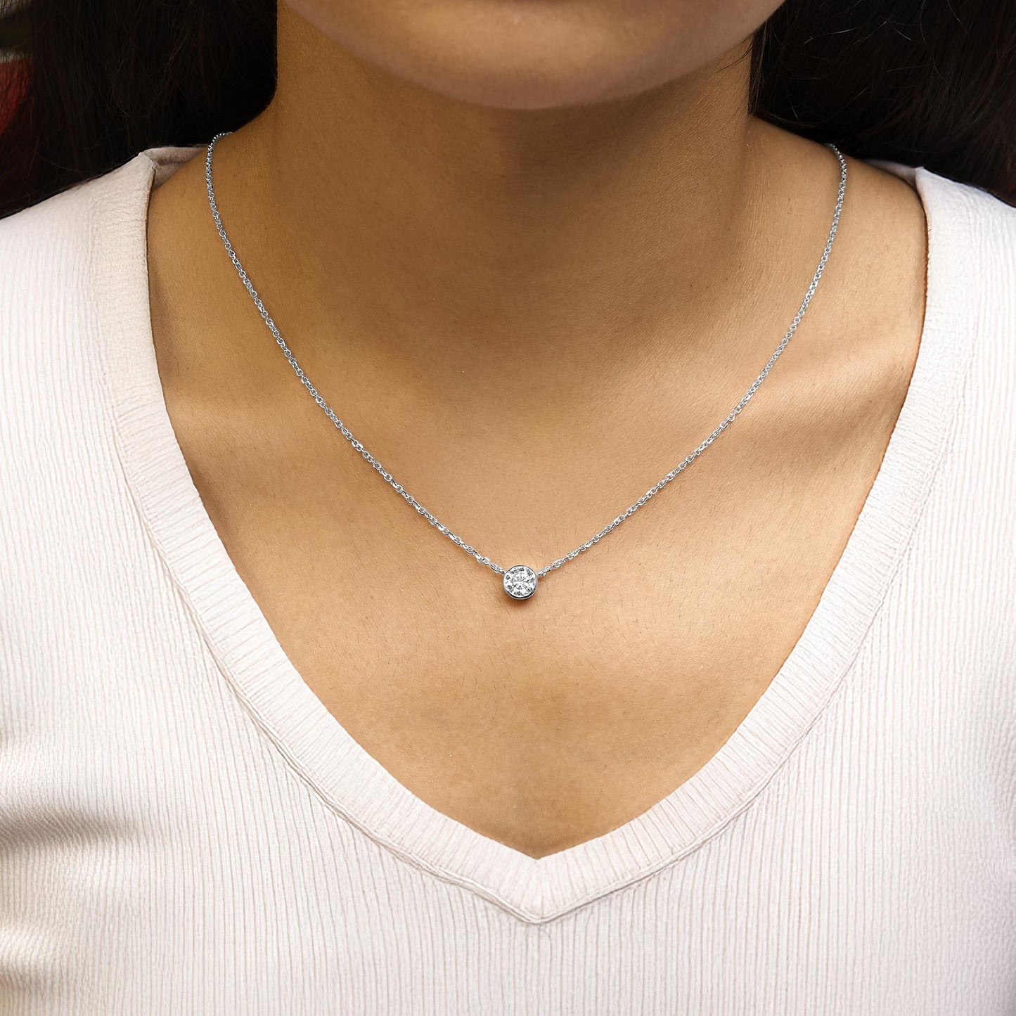 Close-up front view of a white metal necklace with a round bezel-set white gemstone pendant worn on a model.