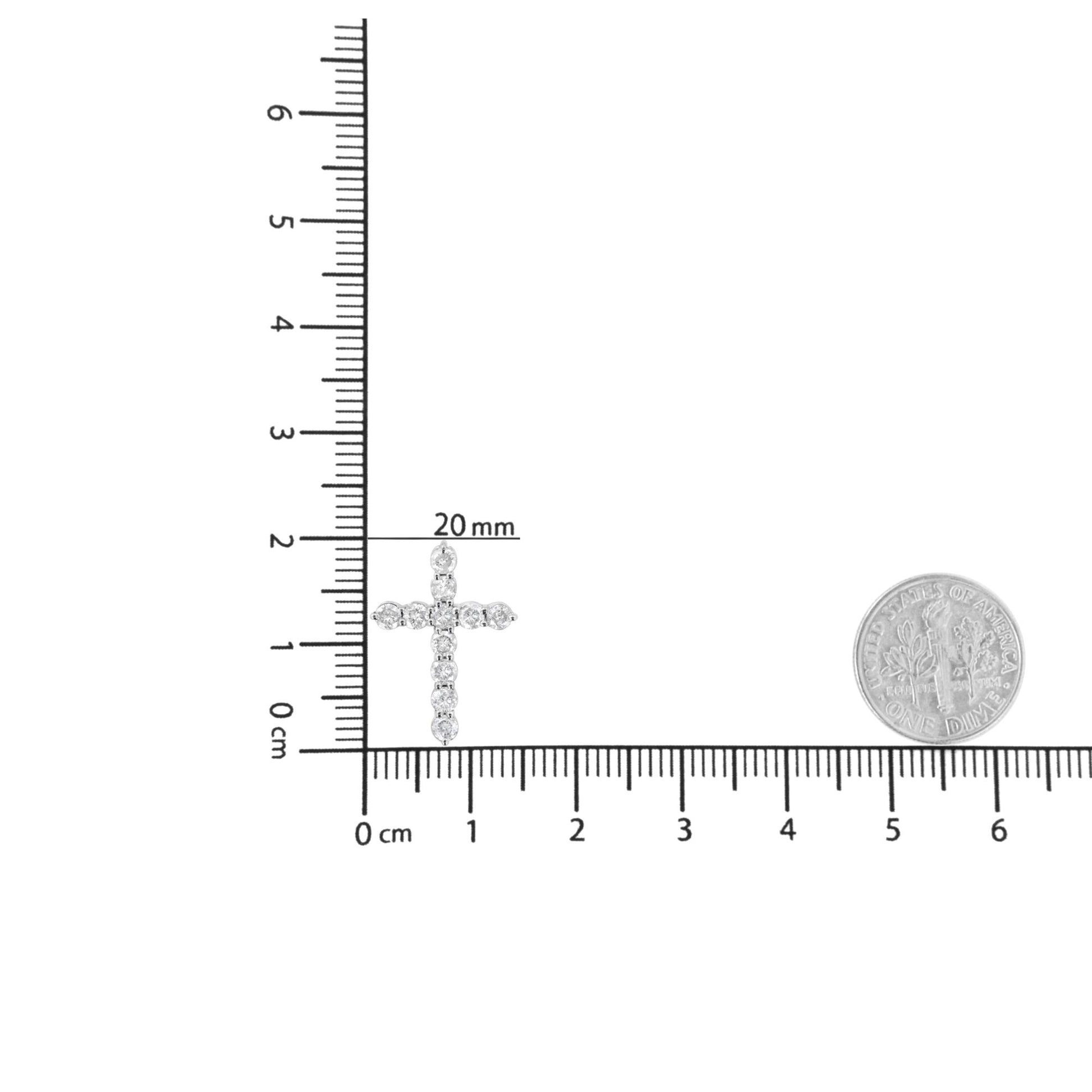 White metal cross-shaped necklace pendant with round white gemstones in prong setting, shown flat on a ruler for size reference with a coin nearby, top-down angle.
