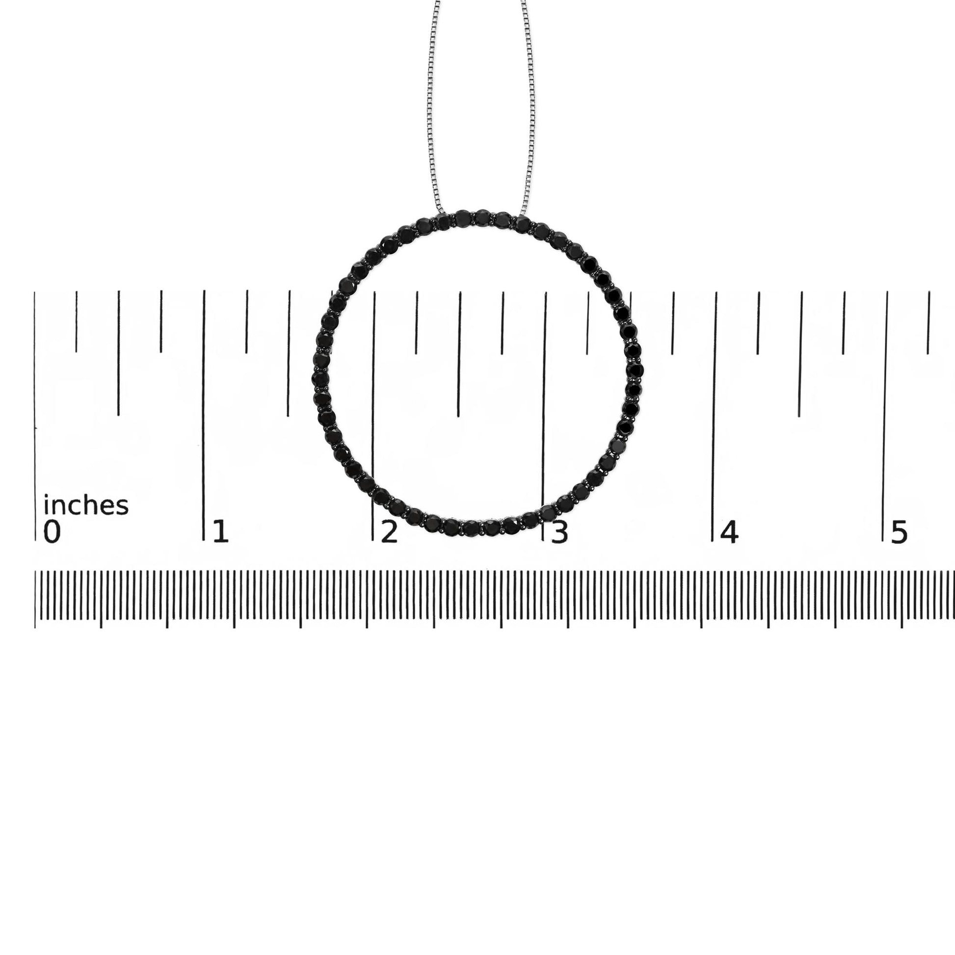 Front-facing shot of a white metal circular necklace pendant set with round black gemstones in a shared prong setting, displayed on a ruler for size reference.