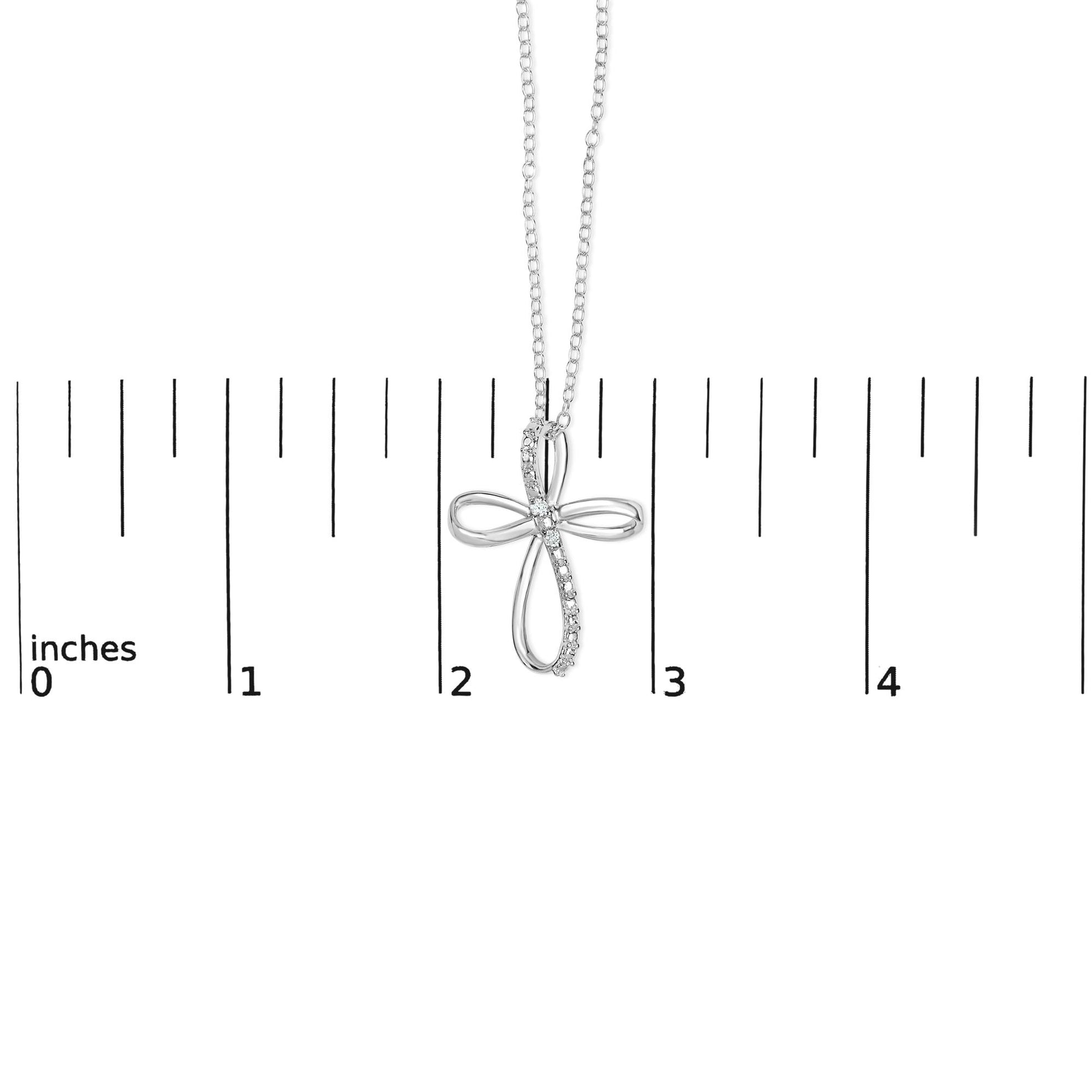 White metal cross-shaped necklace pendant with round white gemstones in prong settings, shown hanging against a ruler for size reference, front-facing angle.