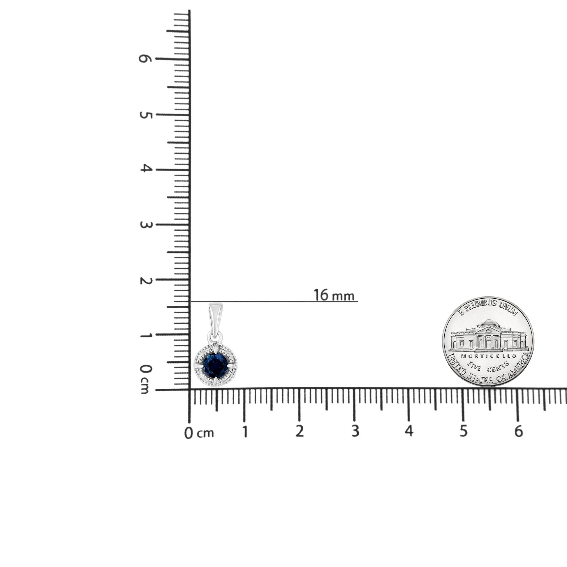 Top-down view of a white metal round pendant necklace with a blue gemstone in a four-prong setting shown on a ruler and next to a coin for size reference.