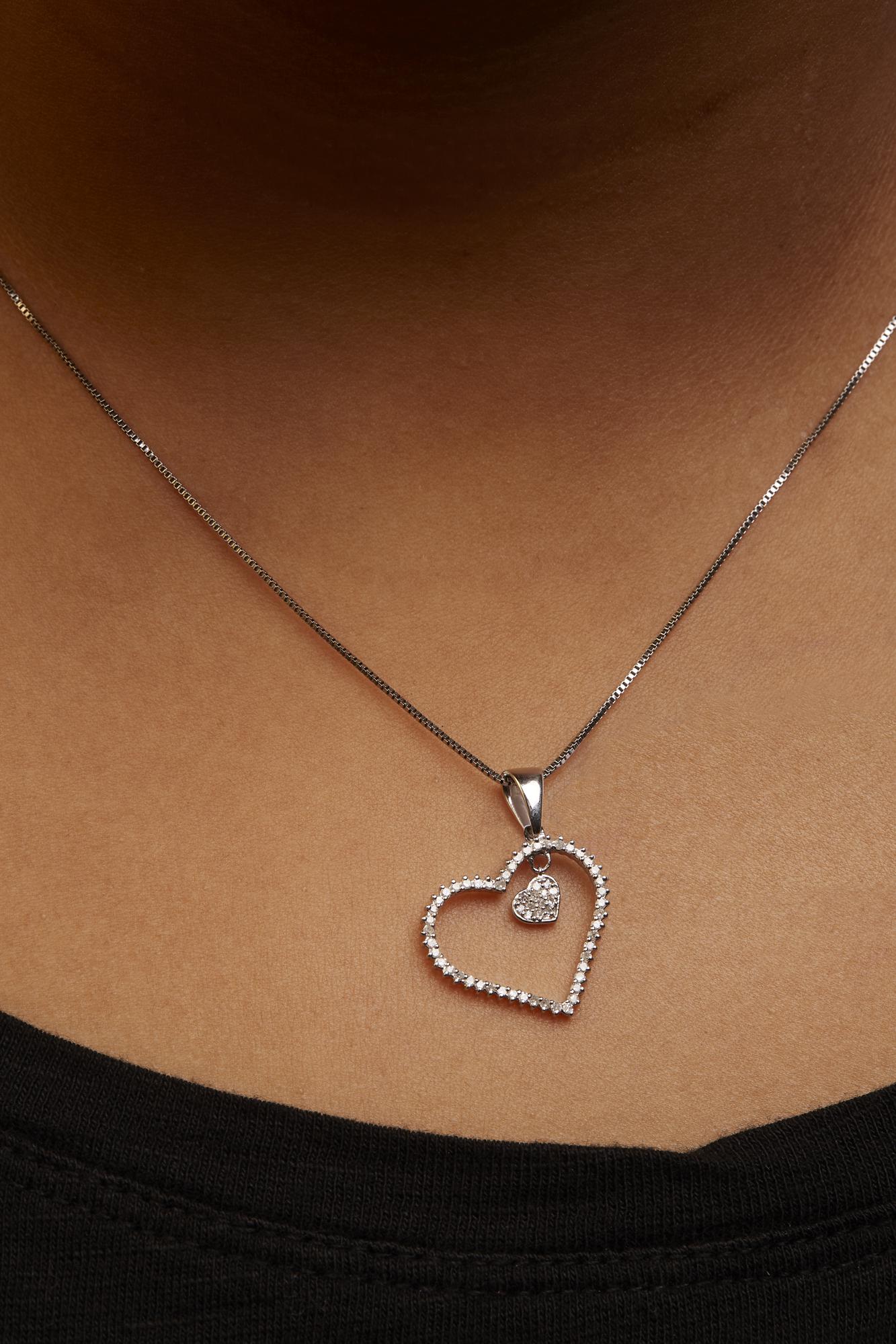 White metal heart-shaped necklace pendant with prong-set round white gemstones around its edge and a smaller heart with white gemstones hanging inside, shown worn on a model, shot from the front at close range.
