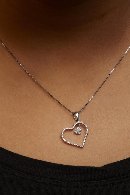 White metal heart-shaped necklace pendant with prong-set round white gemstones around its edge and a smaller heart with white gemstones hanging inside, shown worn on a model, shot from the front at close range.