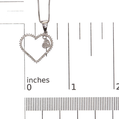 Close-up front angle of a white metal necklace pendant with a heart-shaped open frame adorned with round white gemstones, featuring a smaller solid heart charm covered in round white gemstones, shown on a scale.