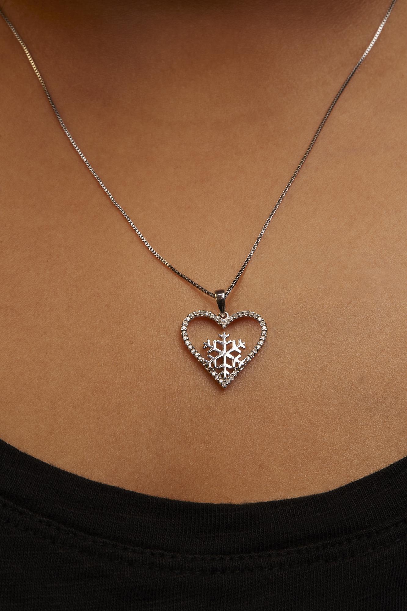 Close-up front view of a white metal necklace with a heart-shaped pendant encrusted with white round gemstones in prong settings, worn on a model. The pendant features a snowflake design inside the heart.