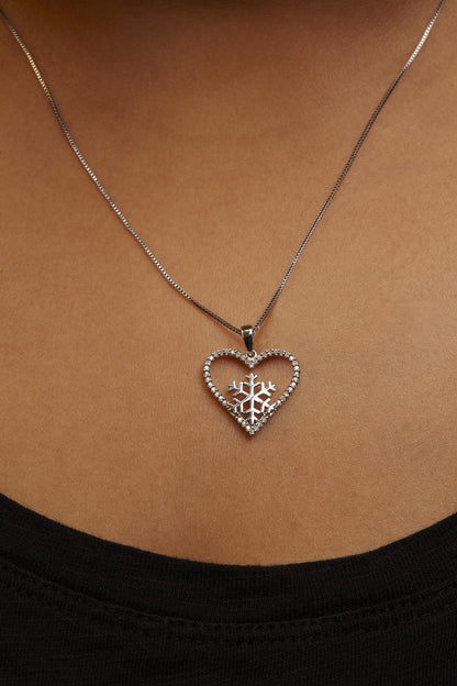 Close-up front view of a white metal necklace with a heart-shaped pendant encrusted with white round gemstones in prong settings, worn on a model. The pendant features a snowflake design inside the heart.
