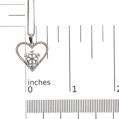 Close-up image of a white necklace pendant with a heart shape adorned with white gemstones set in prongs along the edge and a snowflake design inside, shown on a scale.