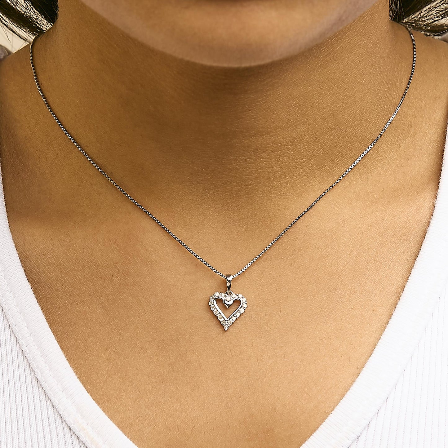 Close-up front view of a white metal heart-shaped pendant necklace with round white gemstones in prong settings, worn on a model.