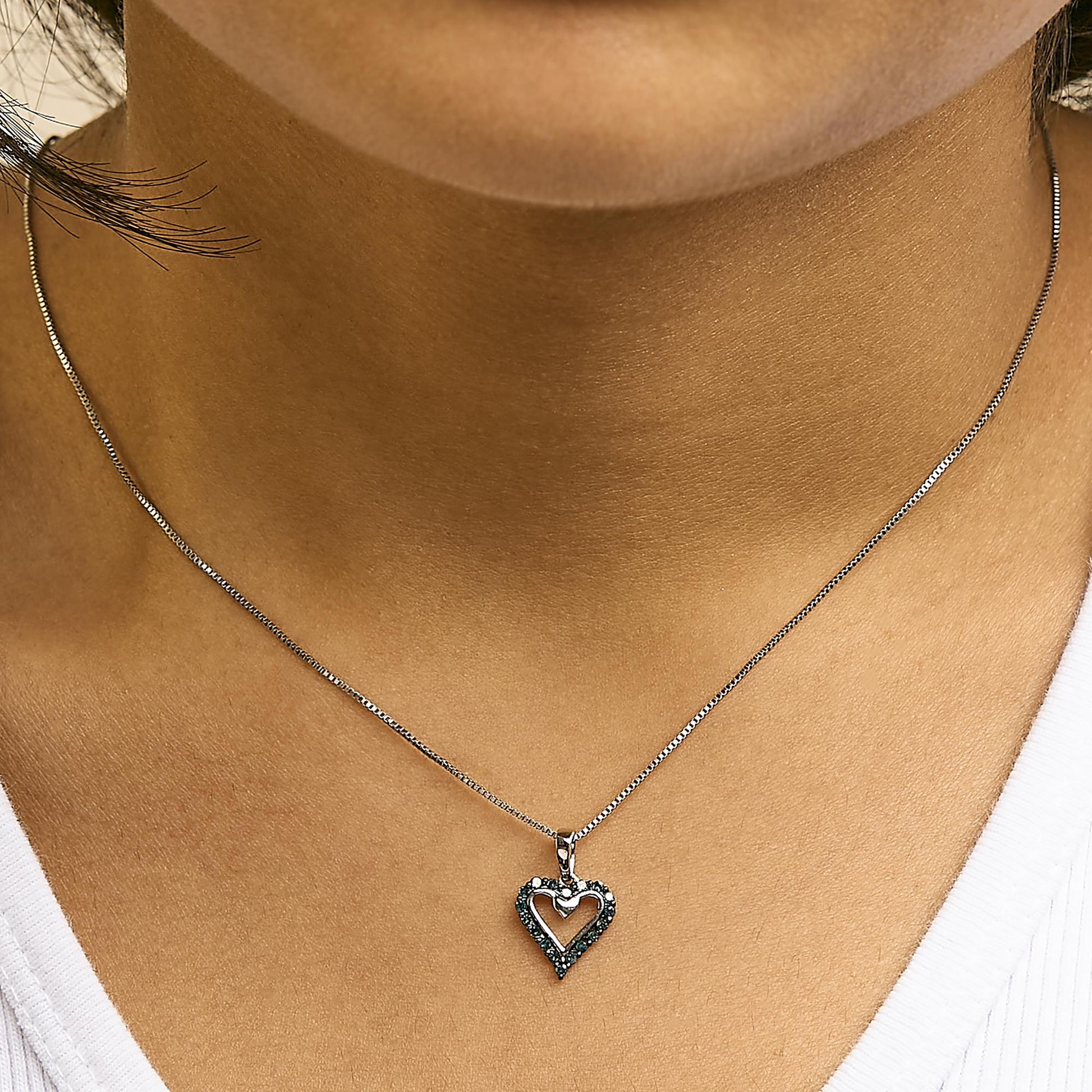 Close-up front view of a white metal necklace with a heart-shaped pendant outlined with round blue gemstones, worn on a model dressed in a dark green shirt.