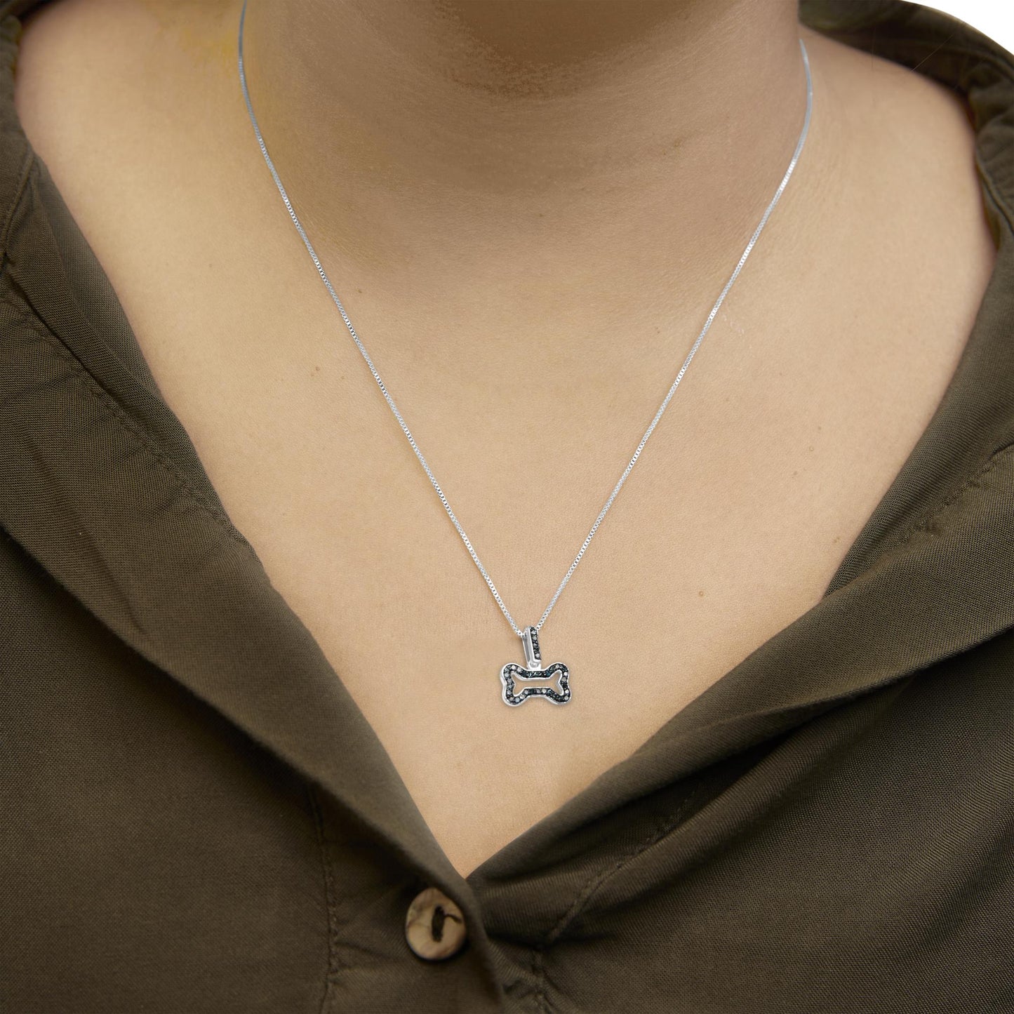 Close-up front view of a white necklace with a bone-shaped pendant adorned with blue round prong-set gemstones, worn on a model with an olive green shirt.