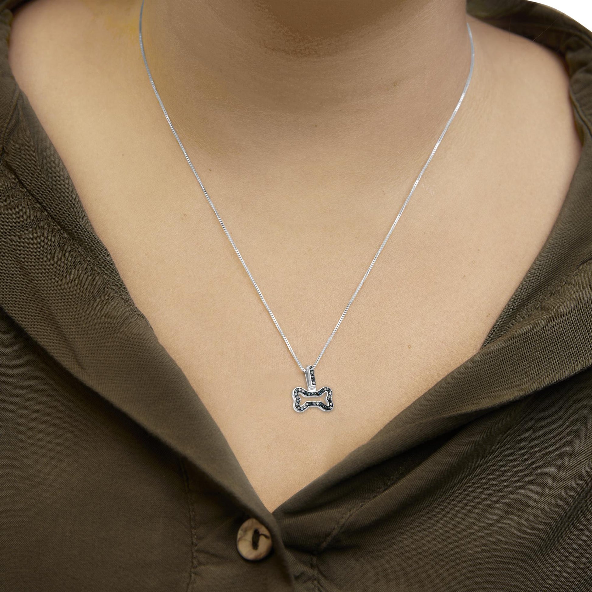 Close-up front view of a white necklace with a bone-shaped pendant adorned with blue round prong-set gemstones, worn on a model with an olive green shirt.