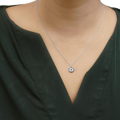 Front view of a white metal necklace with a circular pendant featuring one blue gemstone in the center surrounded by multiple smaller blue gemstones, worn on a model dressed in dark green fabric.