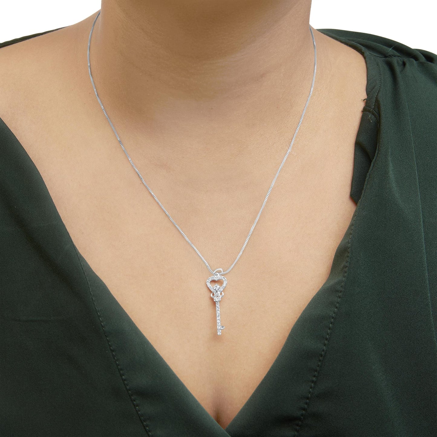 Front-facing close-up of a white metal key-shaped necklace with a heart-shaped top, accented with white round gemstones, worn on a model with a dark green top.