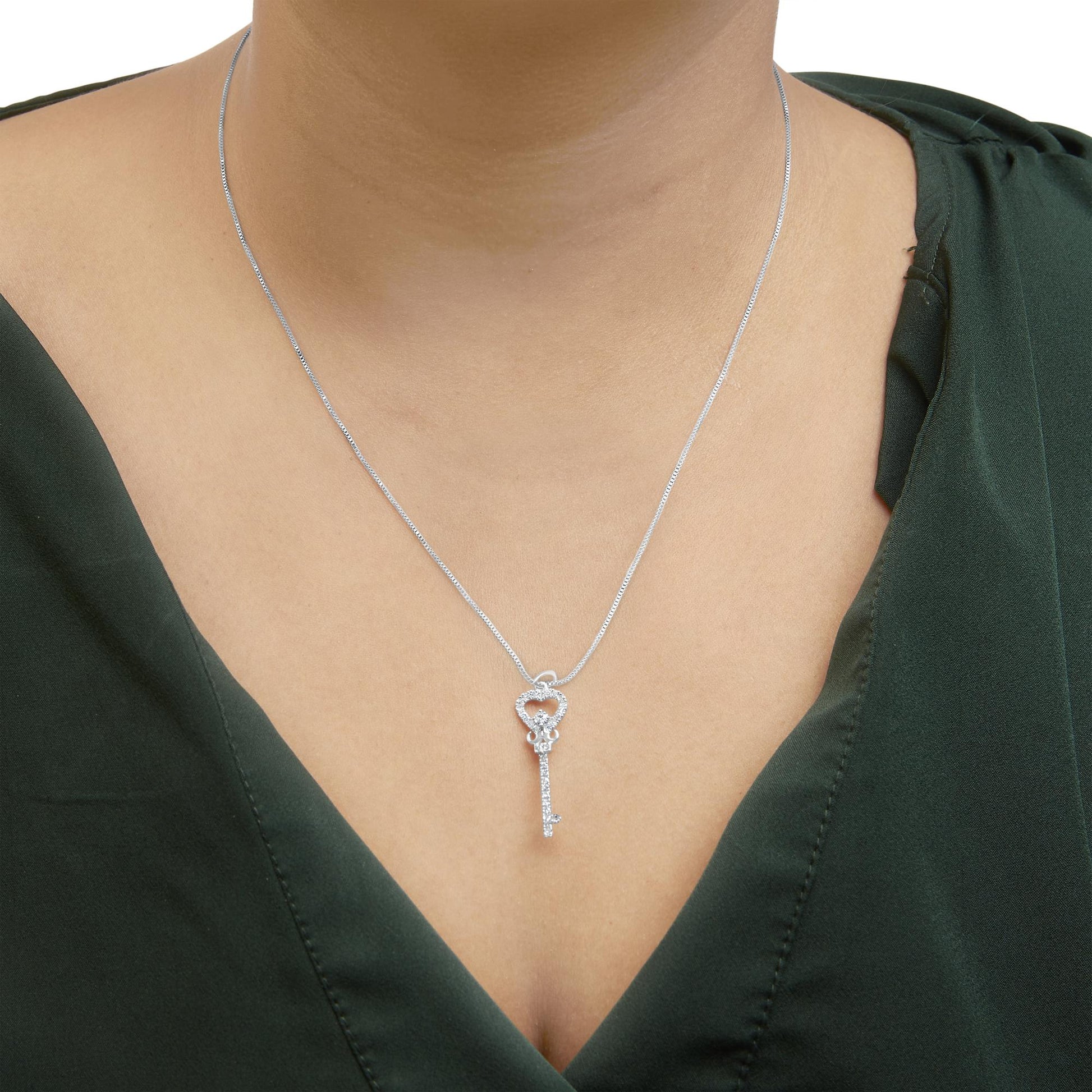 Front-facing close-up of a white metal key-shaped necklace with a heart-shaped top, accented with white round gemstones, worn on a model with a dark green top.