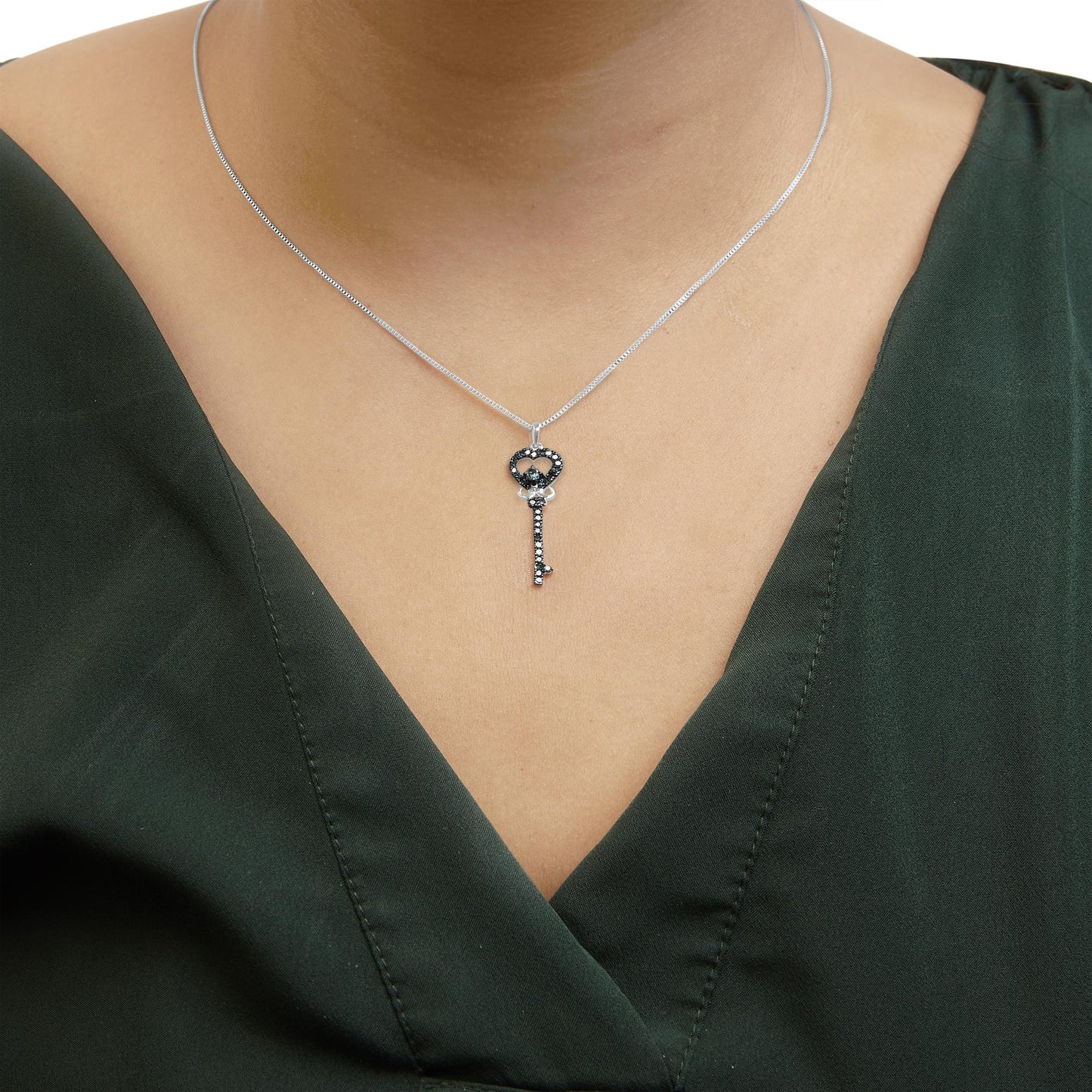 Close-up front view of a necklace on a model, featuring a white chain with a key-shaped pendant adorned with multiple small blue gemstones and one larger blue gemstone in prong settings.