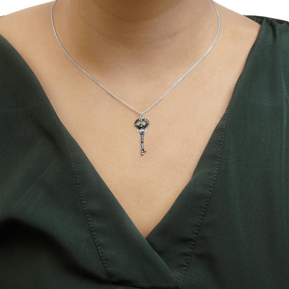 Close-up front view of a necklace on a model, featuring a white chain with a key-shaped pendant adorned with multiple small blue gemstones and one larger blue gemstone in prong settings.