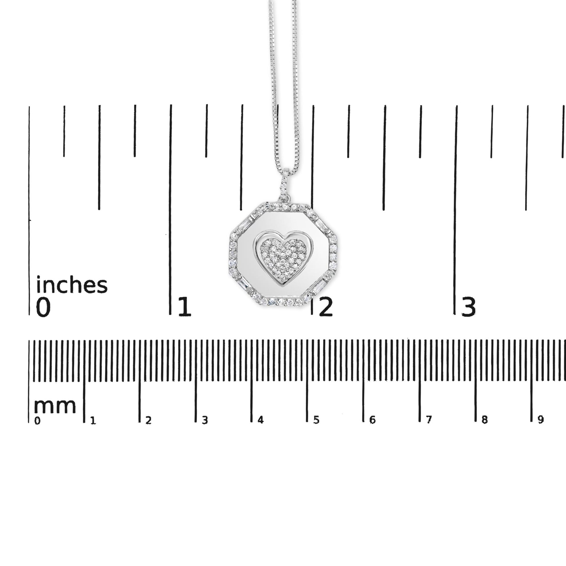 Front view of a white necklace pendant with an octagonal frame set with round and baguette white gemstones surrounding a heart shape filled with round white gemstones, displayed on a ruler for size reference.