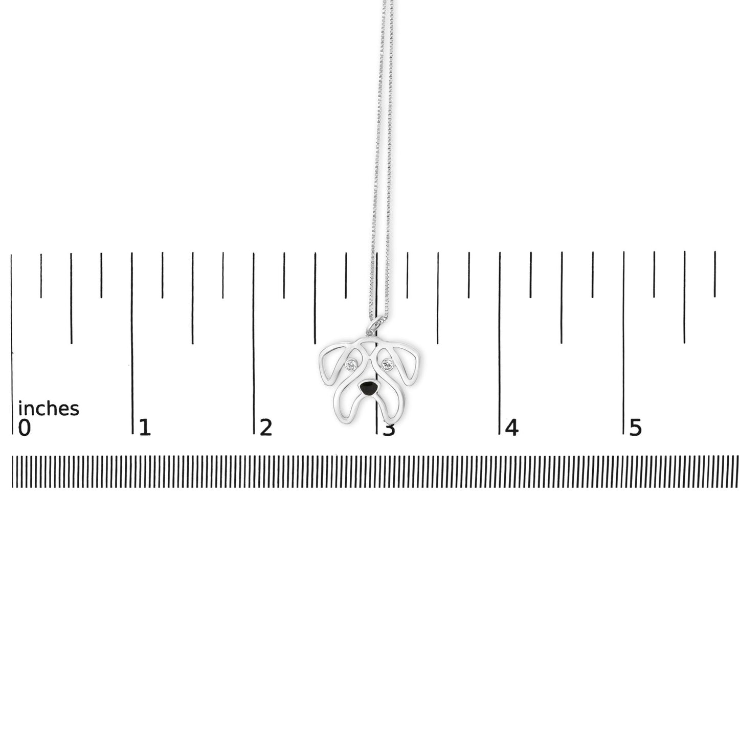 Front-facing shot of a white necklace pendant shaped like a dog’s face with round white gemstones set as eyes, placed on a ruler for size reference.