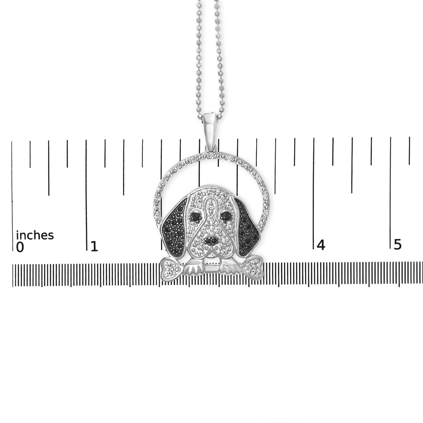 Front view of a white metal necklace pendant shaped like a dog's face with black and white gemstones, resting on a ruler for size reference.