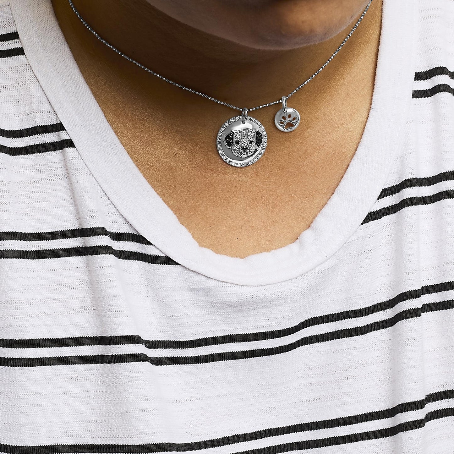 Close-up front view of a white metal necklace worn on a model’s neck with two round pendants, one featuring a dog face adorned with white and black micro-pave gemstones, and a smaller pendant with a cutout paw print.