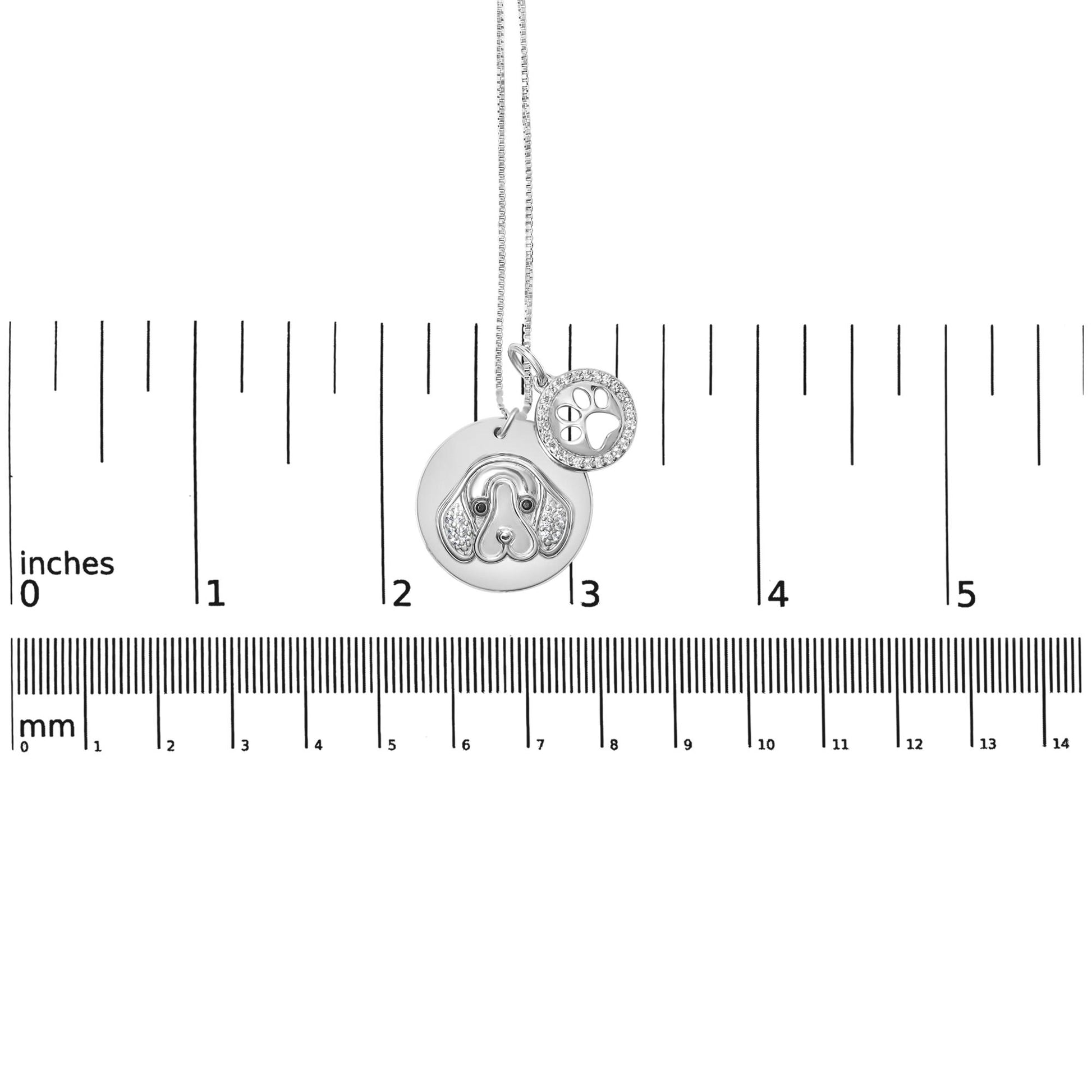 White metal necklace with two round pendants, one featuring a dog face design with white and black gemstones, and the other a paw print surrounded by white gemstones, photographed from the front on a ruler for size reference.