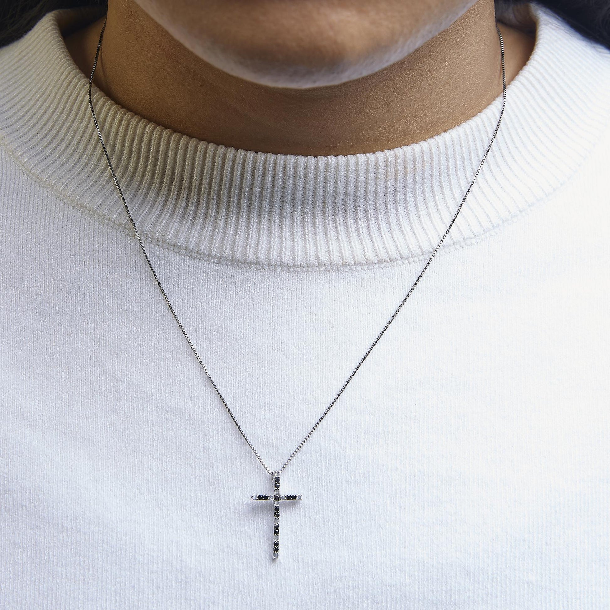 Front view of a white metal cross necklace with alternating black and white round prong-set gemstones, worn on a model with a white ribbed collar sweater.