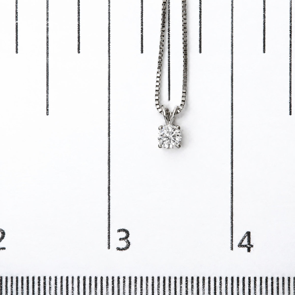 Close-up front view of a white necklace with a round white gemstone in a four-prong setting, shown on a scale with black markings.