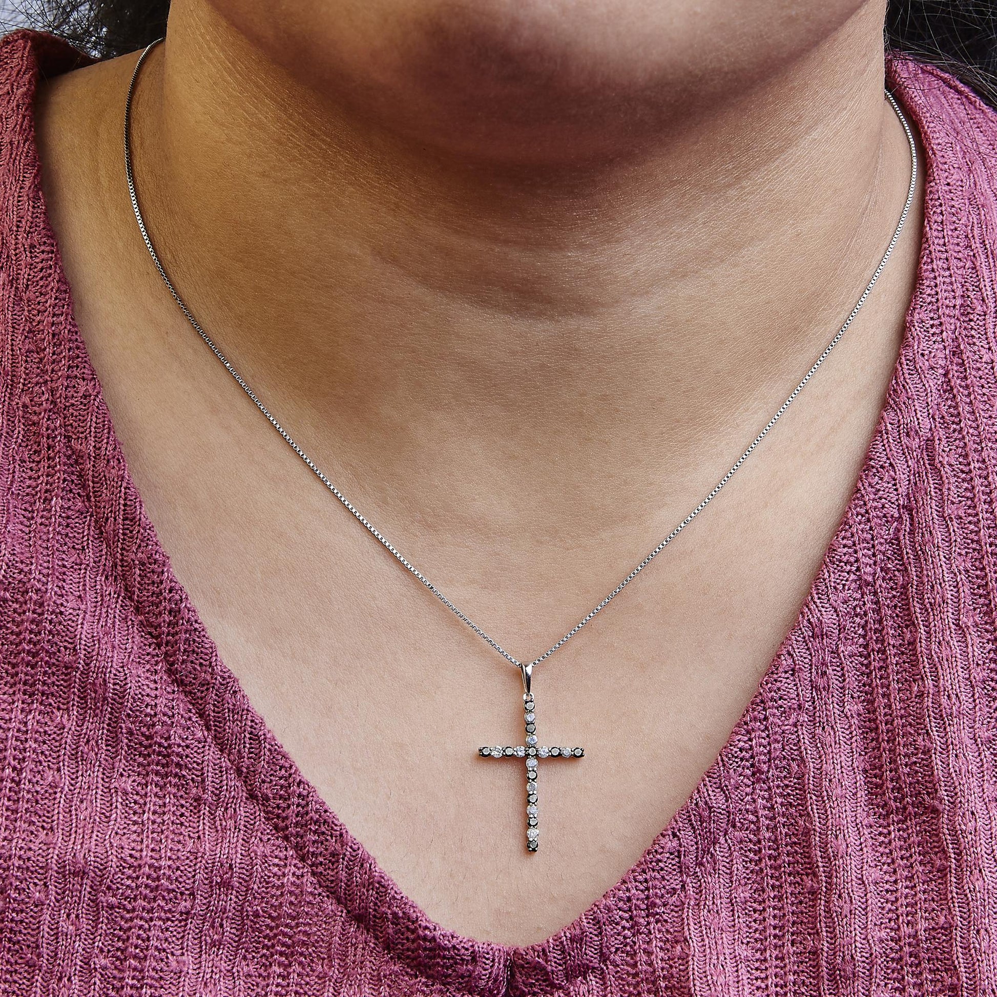 Close-up front view of a necklace on a model featuring a white metal cross pendant adorned with round black and white gemstones in prong settings on a thin chain.