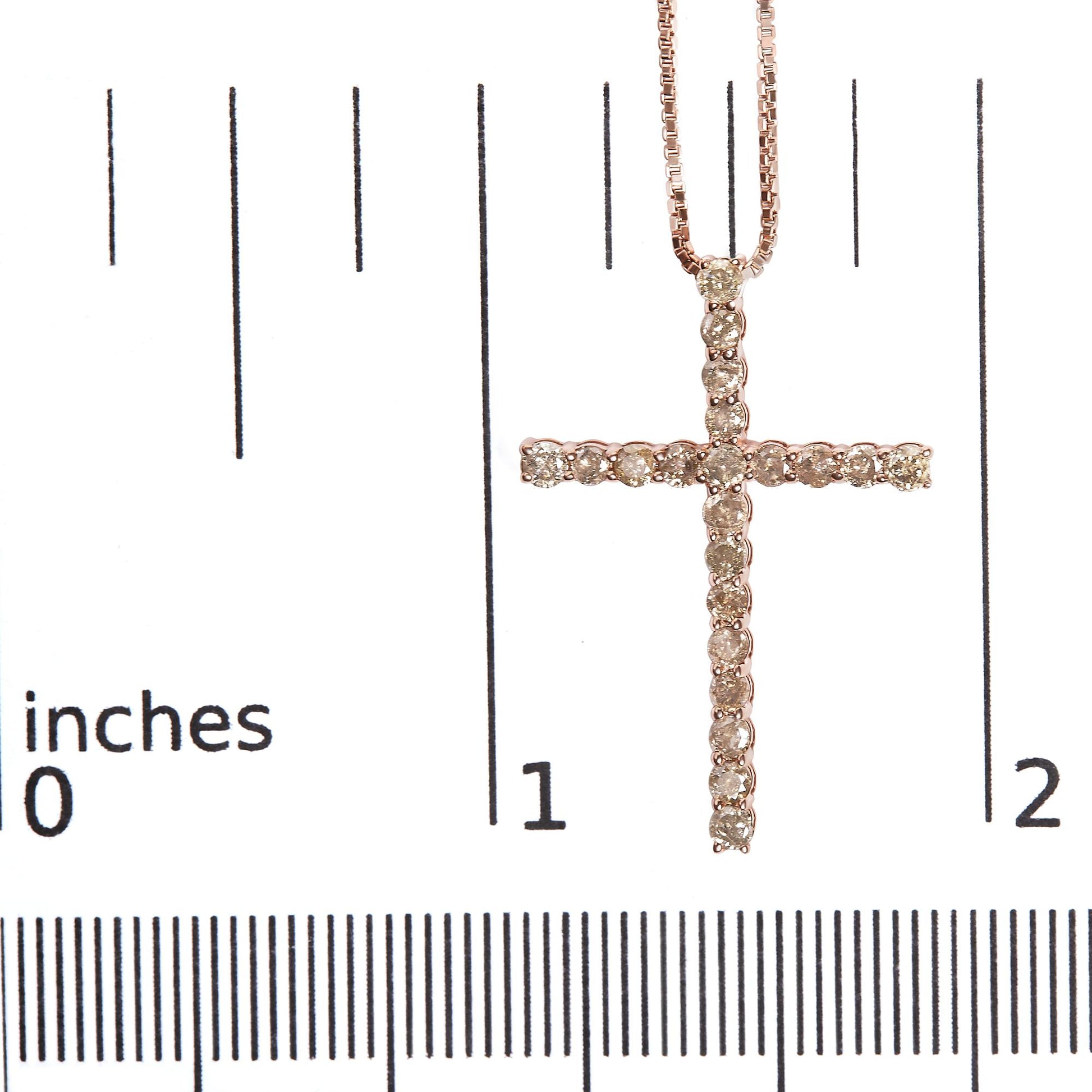 Close-up front view of a rose-colored metal cross necklace pendant set with round champagne-colored gemstones in prong settings, placed on a ruler for size reference.