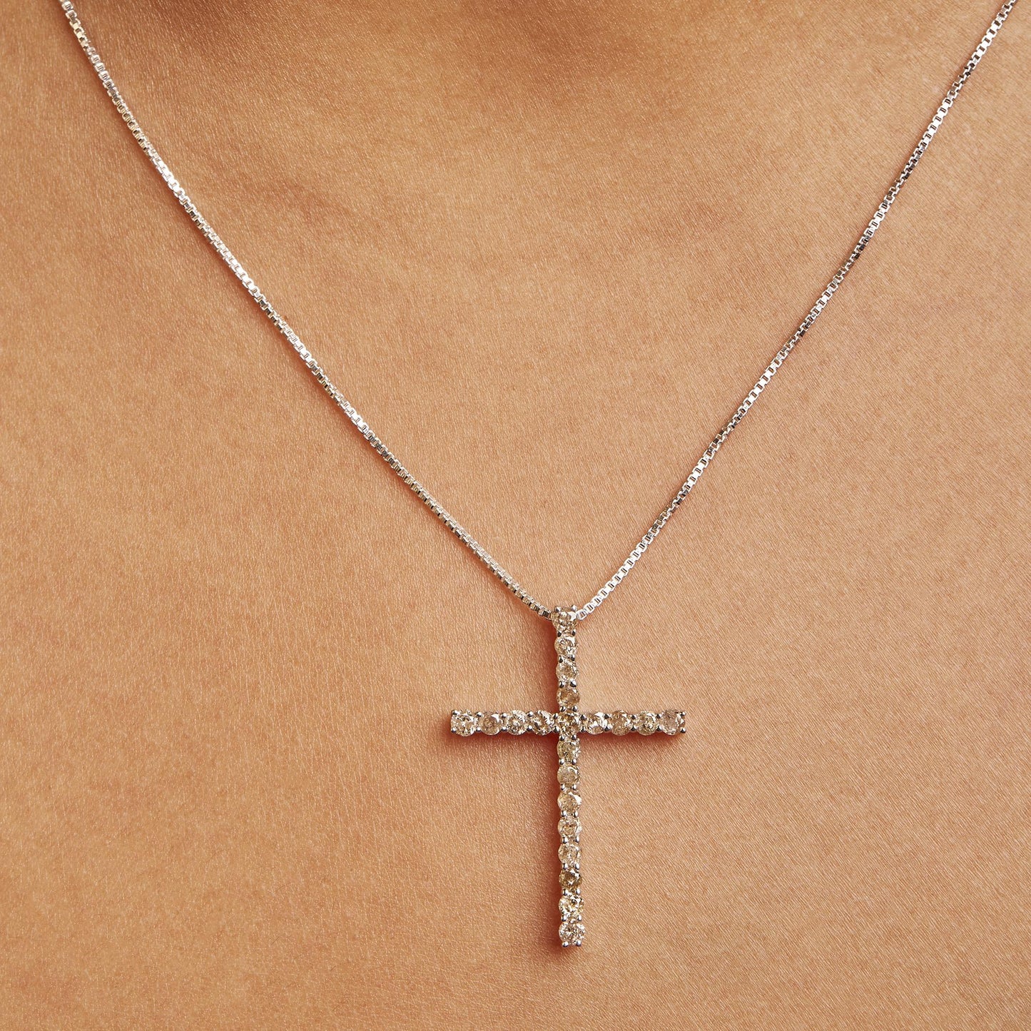 Close-up front view of a white metal necklace with a cross-shaped pendant featuring round champagne-colored gemstones in a prong setting, worn on a model.