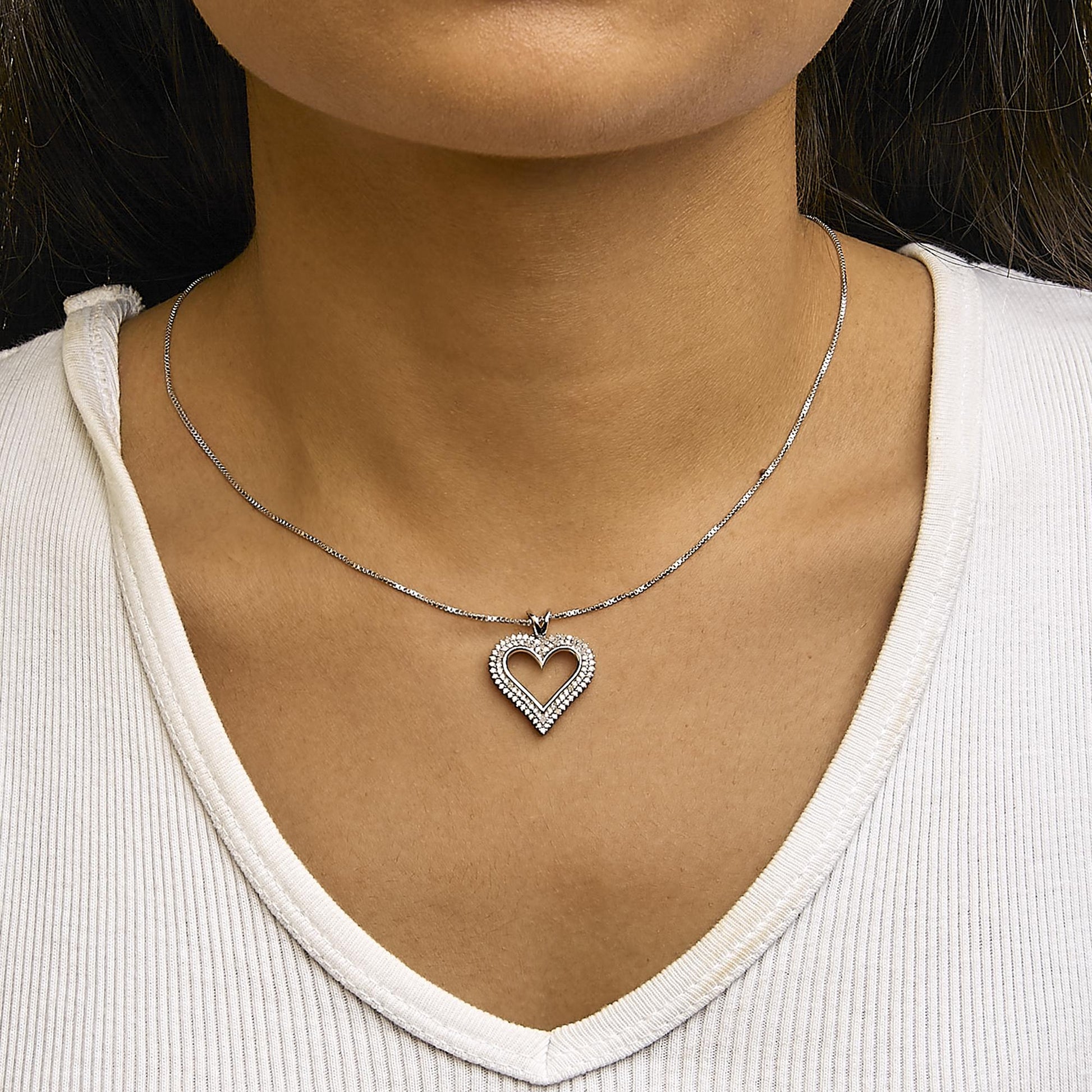 Close-up front view of a white metal necklace worn on a model, featuring a heart-shaped pendant adorned with round white gemstones in prong settings.