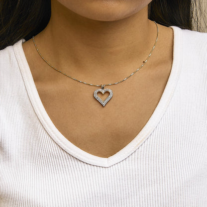 Close-up front view of a heart-shaped yellow metal necklace pendant with round white gemstones set in prongs, worn on a model with a white ribbed top.