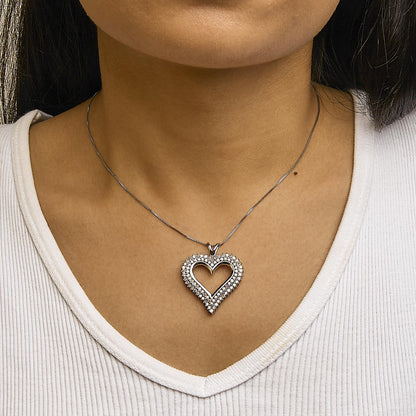 Close-up front view of a white metal heart-shaped necklace pendant encrusted with round white gemstones in prong settings, worn on a model with a white ribbed V-neck top.