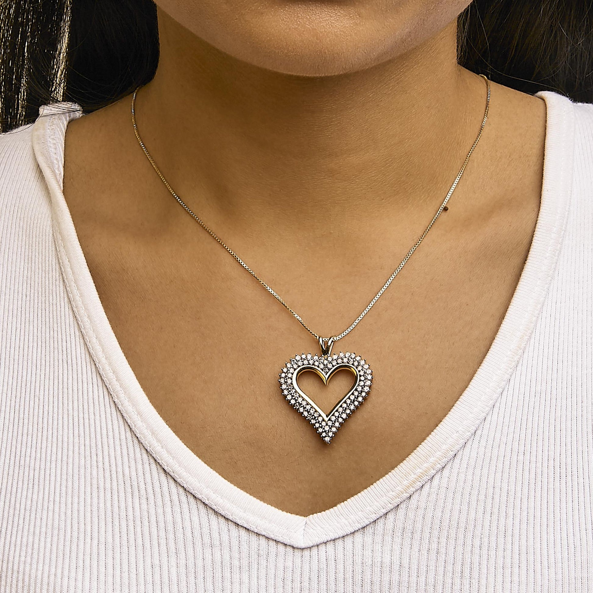 Close-up front view of a yellow metal heart-shaped necklace pendant with two rows of round white gemstones in prong settings, worn on a model.