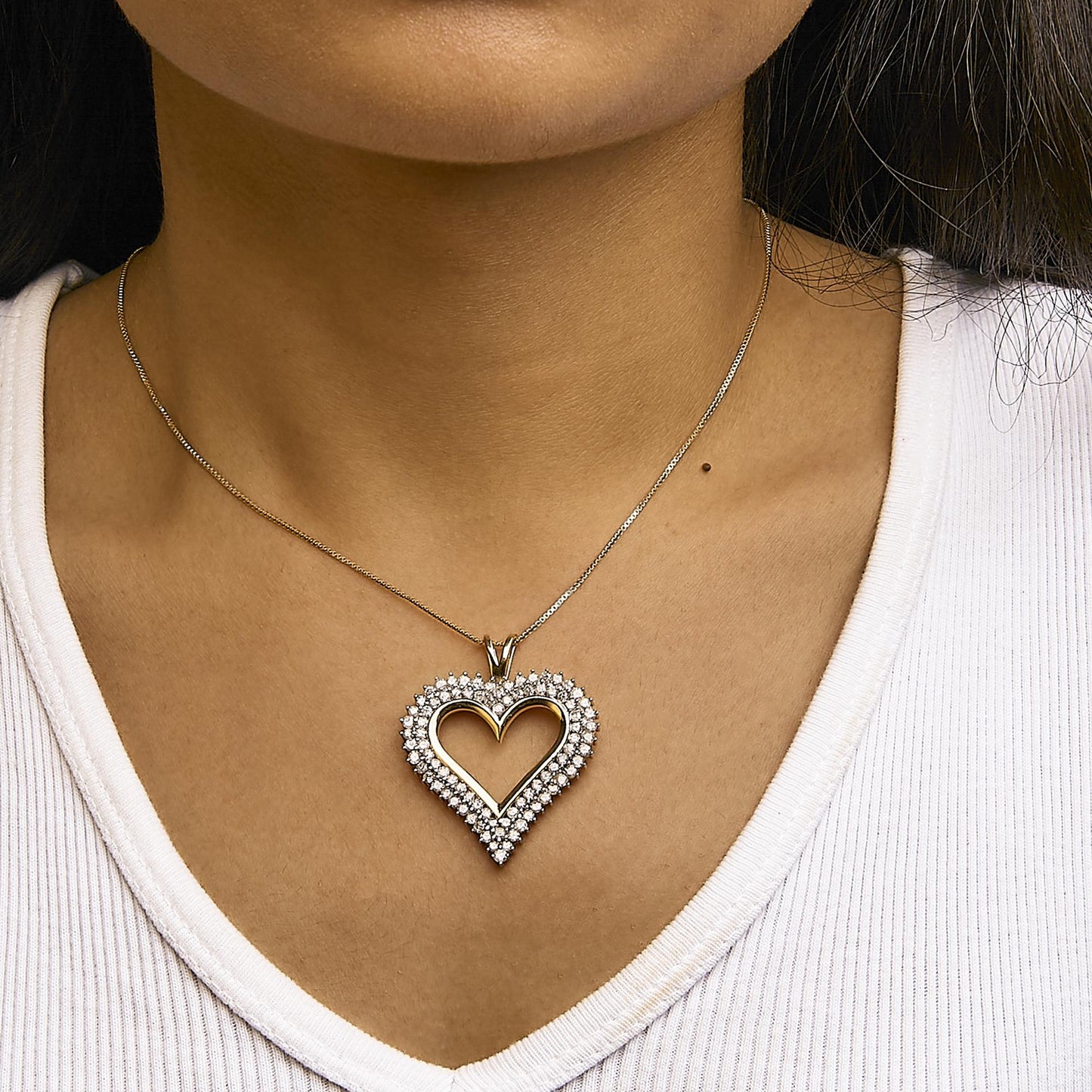 Close-up front view of a yellow metal heart-shaped necklace pendant adorned with two rows of prong-set round white gemstones, worn on a model with a white top.