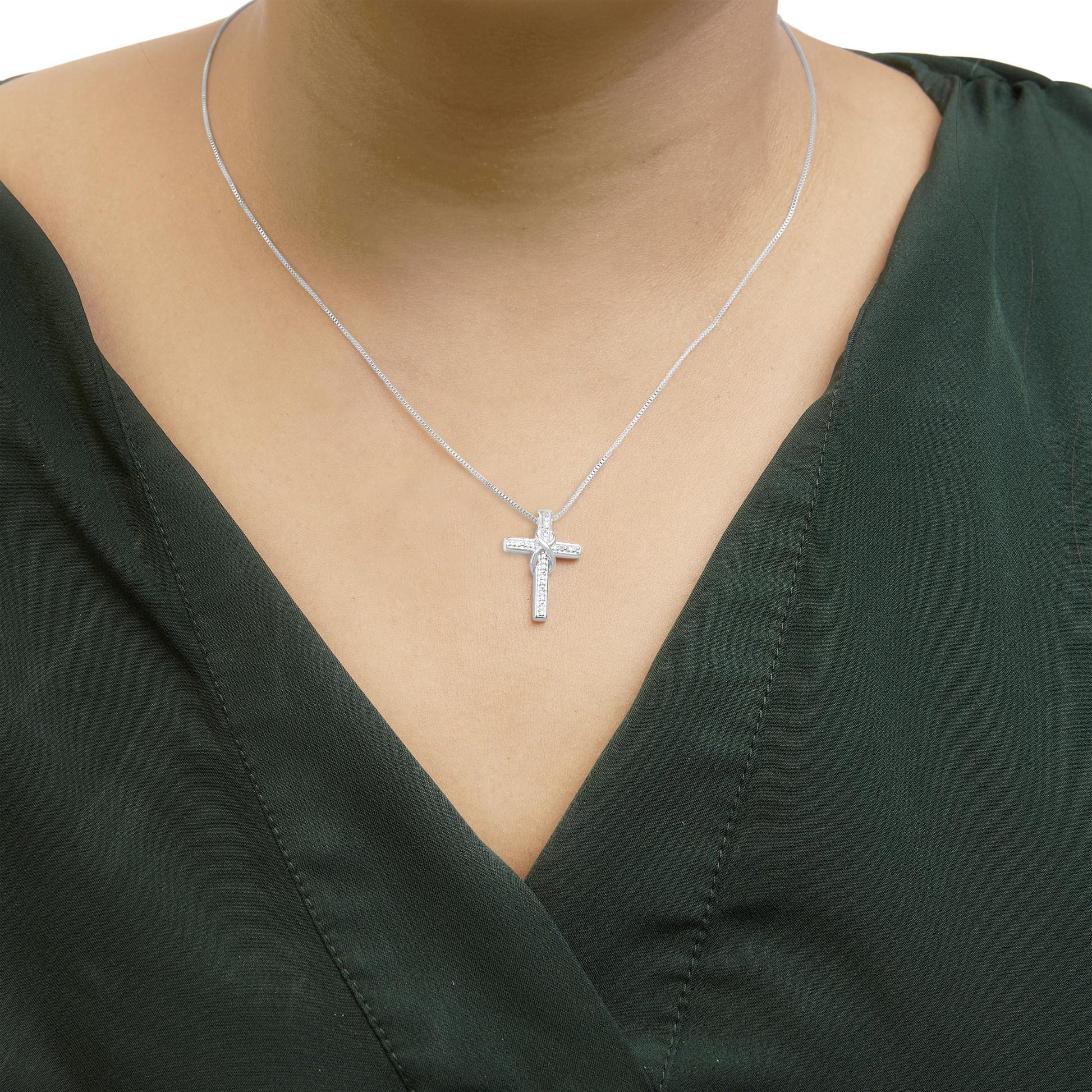 Close-up front view of a white metal necklace with a cross-shaped pendant adorned with round white gemstones worn on a model dressed in a dark green top.