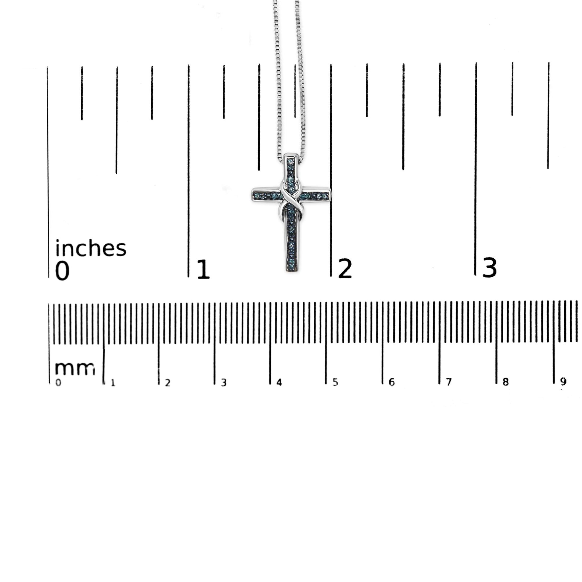 Close-up front view of a white necklace with a cross-shaped pendant adorned with round blue gemstones in a prong setting, displayed on a ruler for size reference.