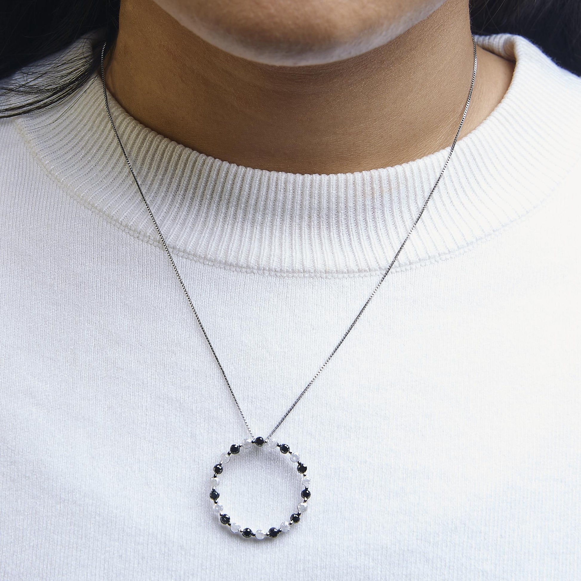 Close-up front view of a white metal necklace on a model, featuring a circular pendant with alternating round black and white gemstones in prong settings.