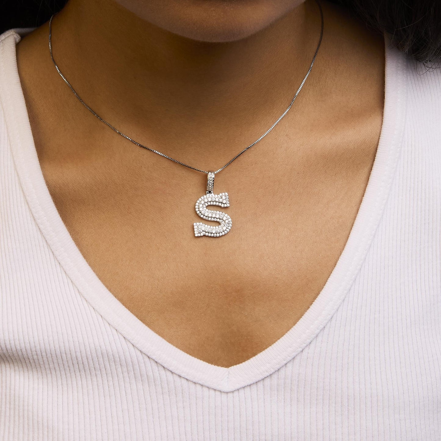 White metal necklace with an 'S' shaped pendant encrusted with round white gemstones in prong settings, shown worn on a model from the front angle.
