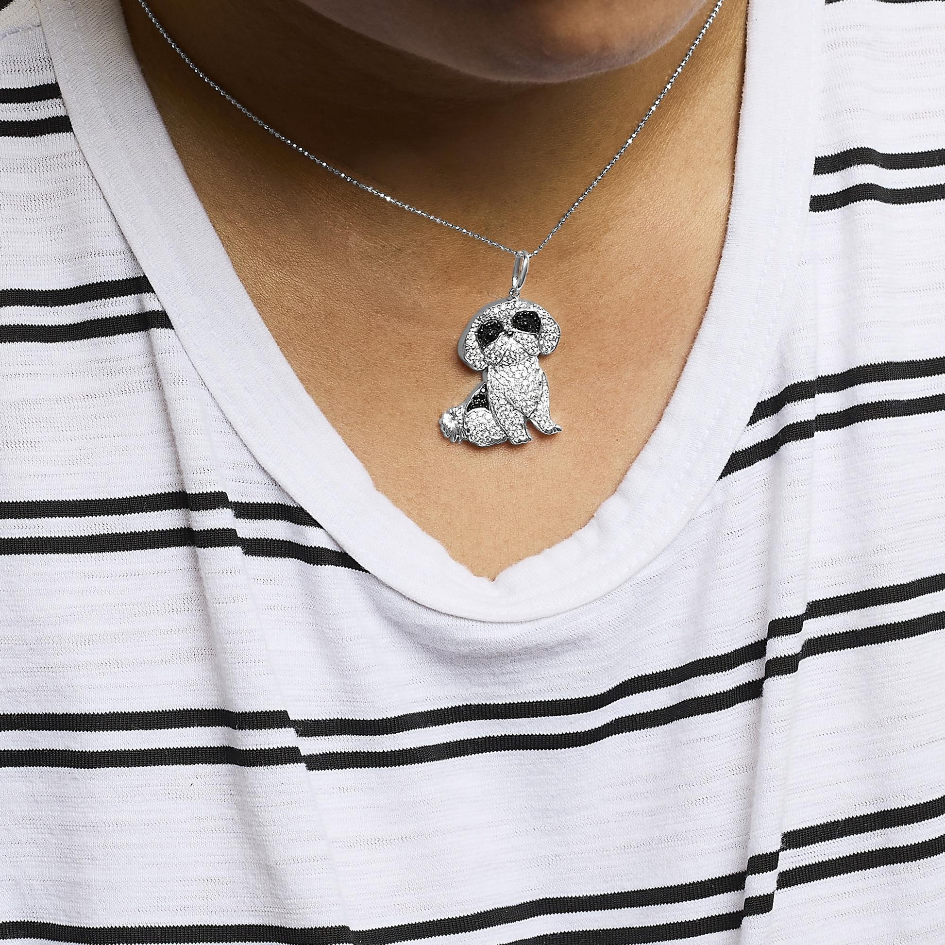 Close-up front shot of a white metal necklace with a dog-shaped pendant adorned with micro-pave set black and white round gemstones, worn on a model with a striped shirt.