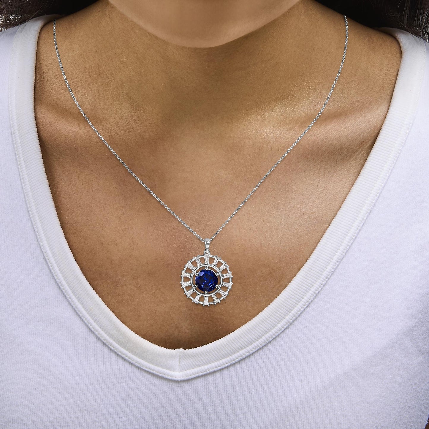 Close-up front view of a model wearing a white metal necklace with a circular pendant featuring a central blue gemstone surrounded by baguette-shaped white gemstones in prong settings.