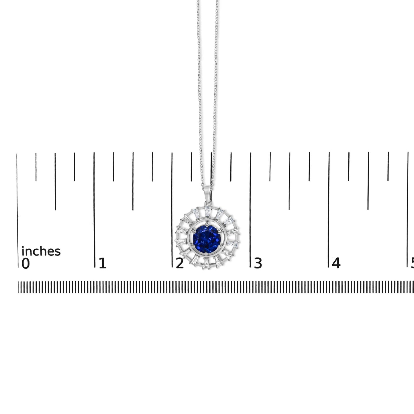 White metal necklace shown on a scale, featuring a circular pendant with a central round blue gemstone surrounded by white baguette-shaped gemstones arranged radially, viewed from a top-down angle.