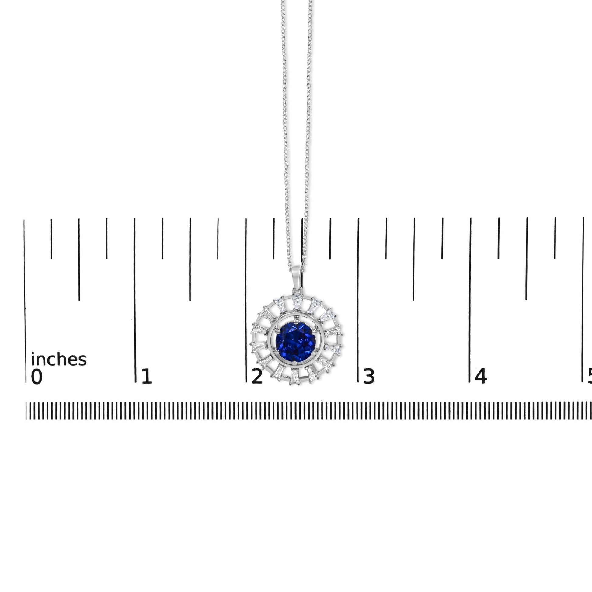 White metal necklace shown on a scale, featuring a circular pendant with a central round blue gemstone surrounded by white baguette-shaped gemstones arranged radially, viewed from a top-down angle.