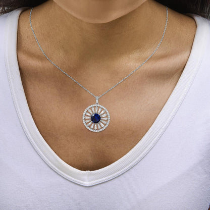 White metal necklace with a circular pendant featuring a large blue gemstone center, surrounded by alternating baguette and round white gemstones, shown worn on a model in a close-up front view.