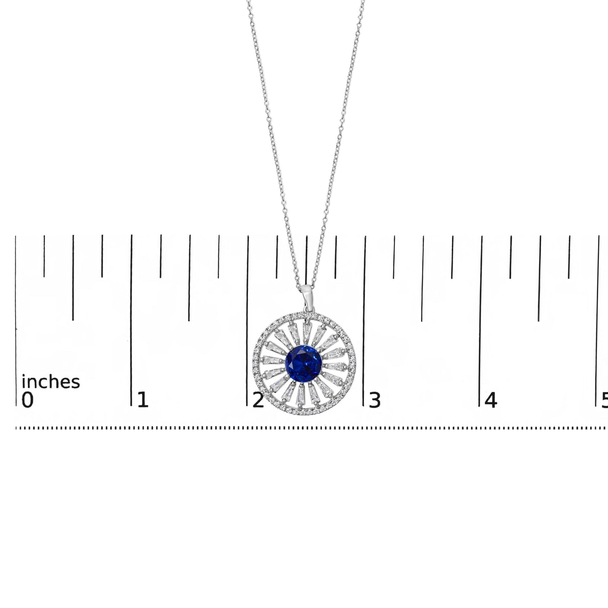 Close-up image of a white metal necklace pendant shown on a scale, featuring a circular design with a central blue gemstone surrounded by alternating baguette and round white gemstones in a radial pattern.