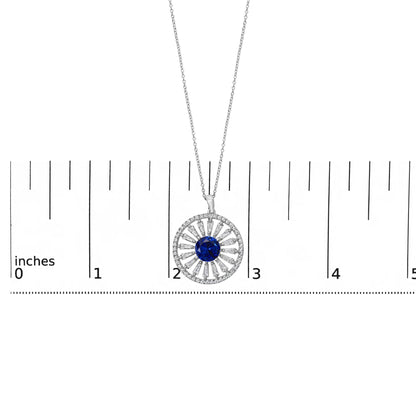 Close-up image of a white metal necklace pendant shown on a scale, featuring a circular design with a central blue gemstone surrounded by alternating baguette and round white gemstones in a radial pattern.