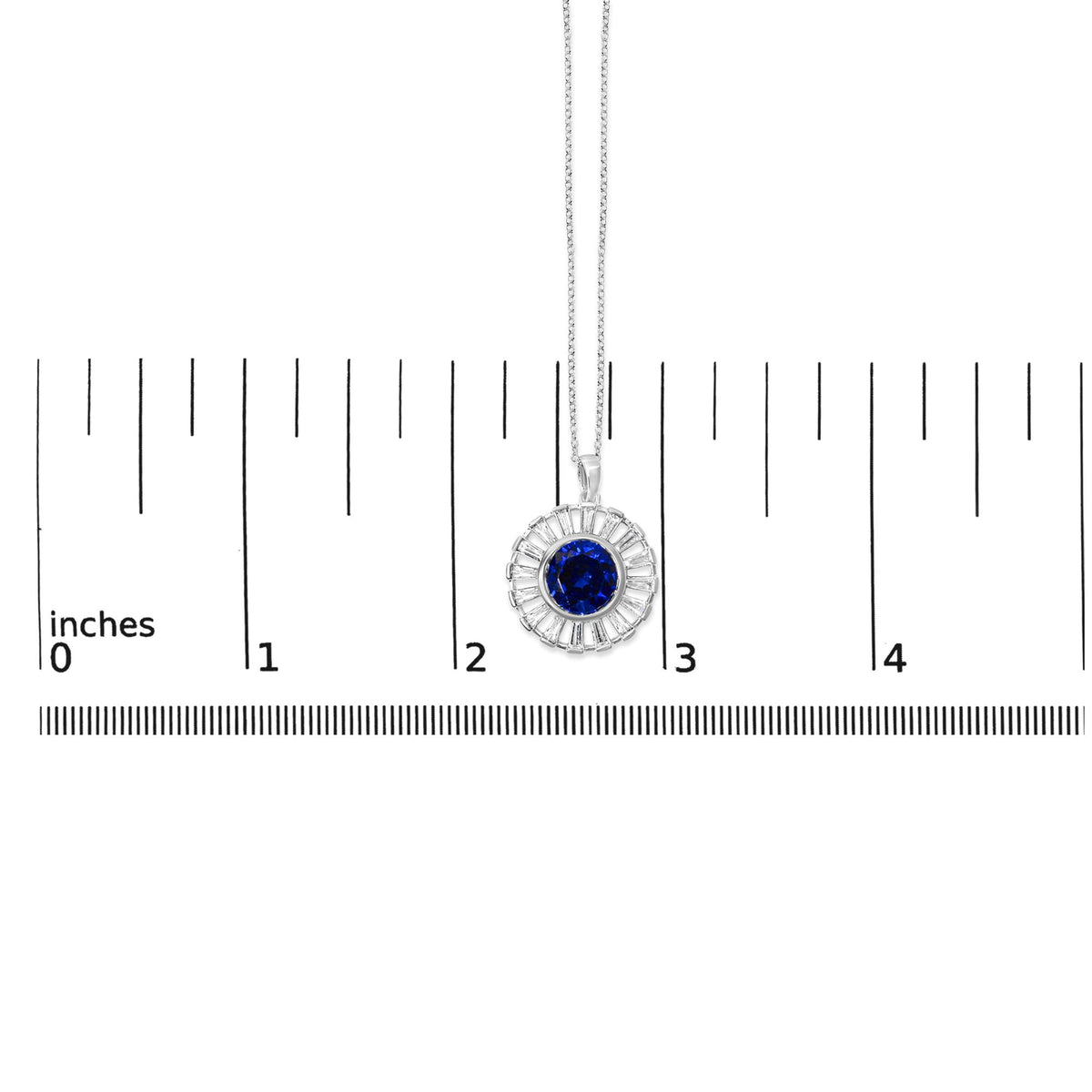 White metal necklace shown on a scale, featuring a circular pendant with a central round blue gemstone surrounded by baguette-shaped white gemstones in a channel setting. Shot is a close-up, front-facing view.