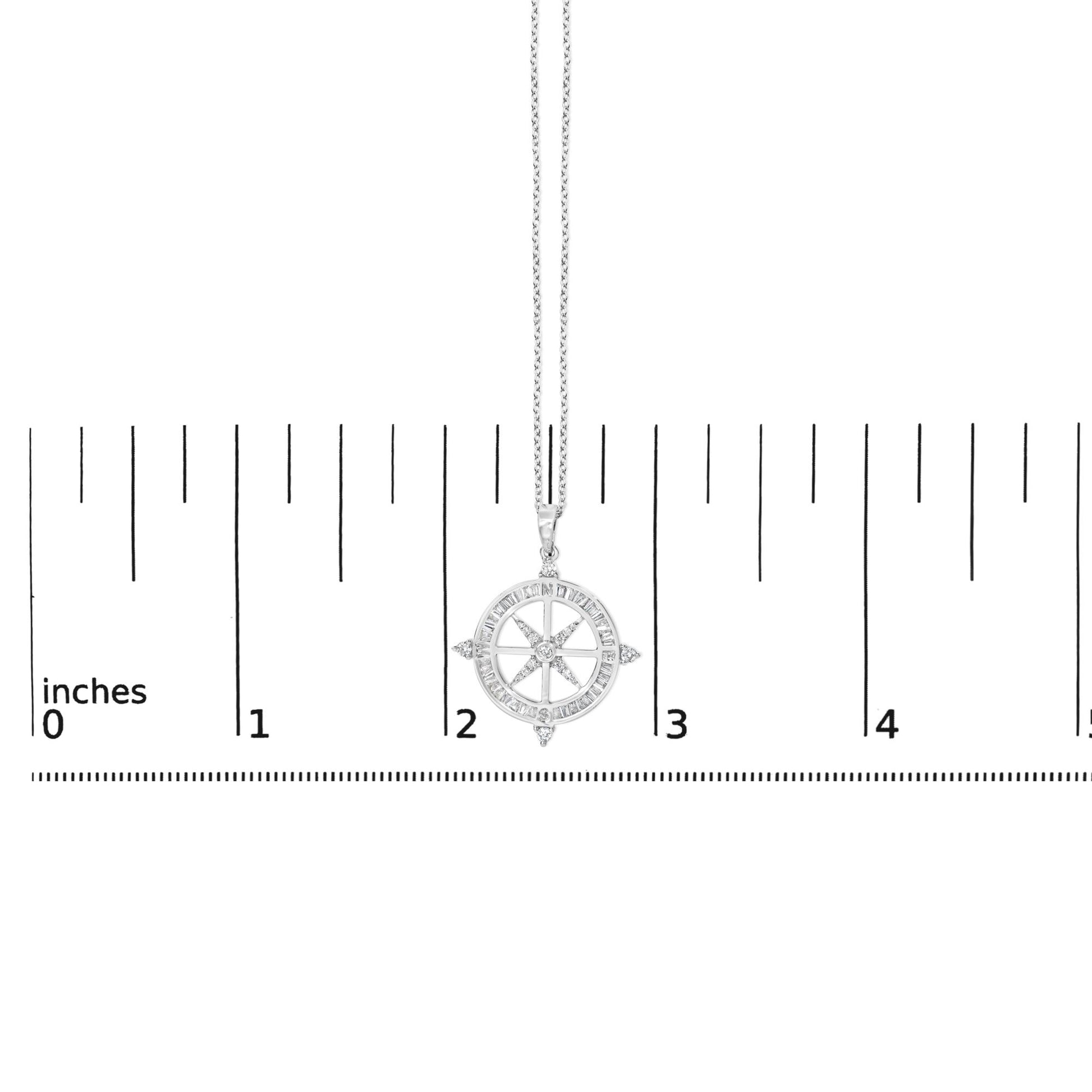 Close-up front view of a white metal necklace with a compass-shaped pendant. The pendant features a circular design with baguette-shaped white gemstones channel set around the outer edge and round white gemstones prong set within the compass points. The necklace is shown on a scale.