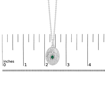 Close-up front view of a white metal necklace pendant with an oval shape, featuring a central green oval gemstone surrounded by white baguette and round gemstones, shown on a scale.